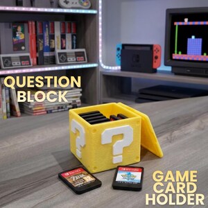 Op de afbeelding: Een gele spelkaarthouder in de vorm van een vraagtekenblok met witte vraagtekens. Twee spelkaarten liggen voor de houder. De tekst "QUESTION BLOCK" en "GAME CARD HOLDER" staat in de afbeelding.