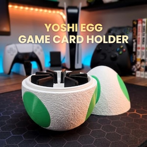 Może przedstawiać: Uchwyt na karty do gier Yoshi Egg, biały z zielonymi akcentami, o teksturowanej powierzchni. Uchwyt jest otwarty, ukazując czarne karty do gier. Design nawiązuje do jaja Yoshiego.