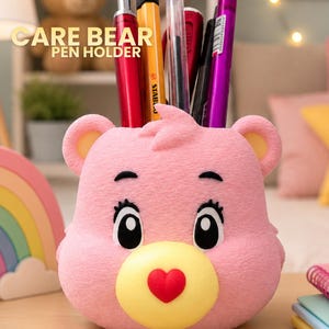 Könnte beinhalten: Ein rosa Stiftehalter in Bärenform mit gelber Schnauze und roter herzförmiger Nase. Der Bär hat schwarze Augen mit langen Wimpern und kleine Ohren. Mehrere Stifte und Marker stecken in der oberen Öffnung. Der Text CARE BEAR PEN HOLDER ist zu sehen.