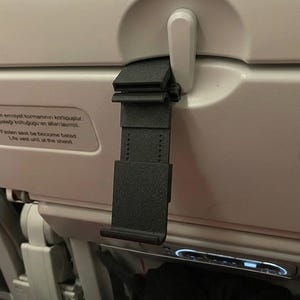 May include: A black, adjustable phone holder attached to the back of an airplane seat. The holder is made of a textured, dark material and is designed to secure a phone or tablet. The seat back is white with safety instructions.