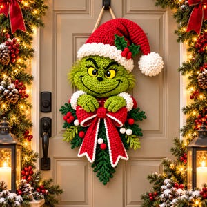 May include: A crocheted wall hanging featuring a green character with yellow eyes and a wide smile, wearing a red and white Santa hat. The character holds a large red bow with white trim, accented by green pine sprigs, red berries, and small white pom-poms.