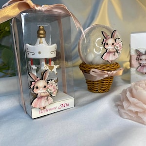 May include: A carousel-themed baby shower favor in a clear box with a pink ribbon. The carousel is white with gold accents and small horse figures. A bunny figure in a pink dress holds flowers. The text "Welcome Mia" is printed on the box.