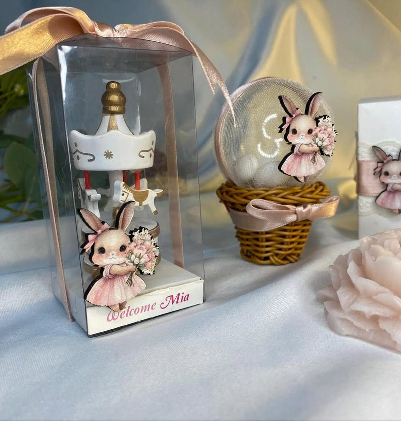 May include: A carousel-themed baby shower favor in a clear box with a pink ribbon. The carousel is white with gold accents and small horse figures. A bunny figure in a pink dress holds flowers. The text "Welcome Mia" is printed on the box.