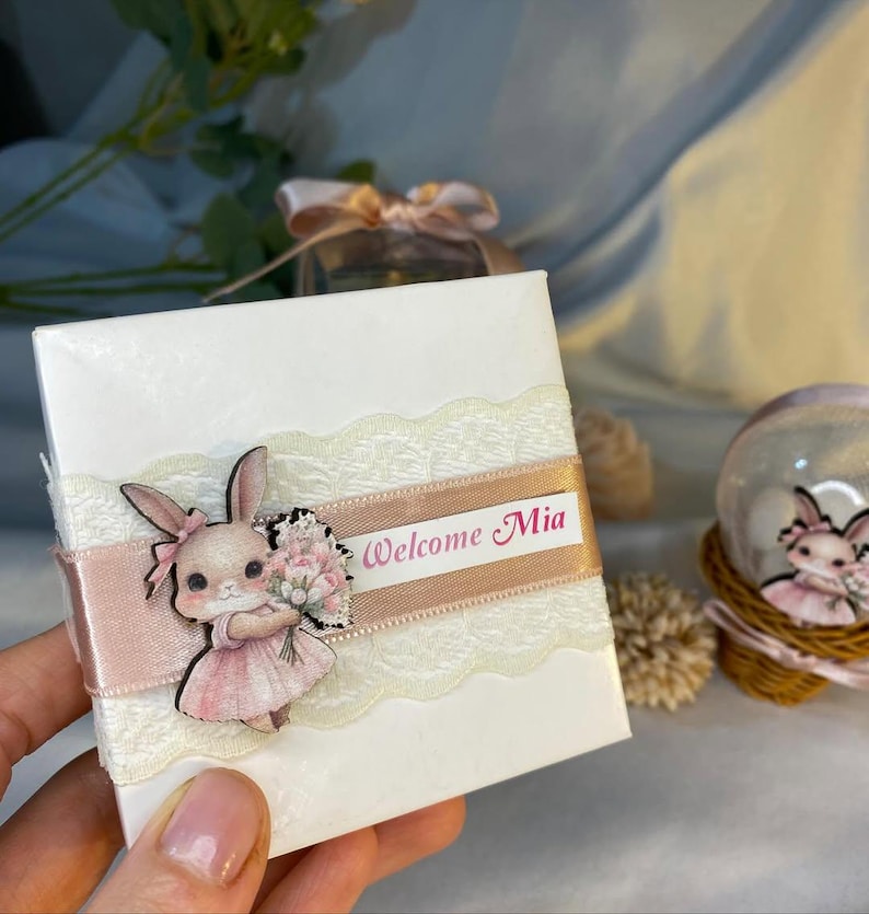 May include: A white gift box decorated with a pink ribbon and lace trim. A wooden bunny illustration in a pink dress is attached to the box. The text "Welcome Mia" is printed on the ribbon. A snow globe with a bunny is in the background.