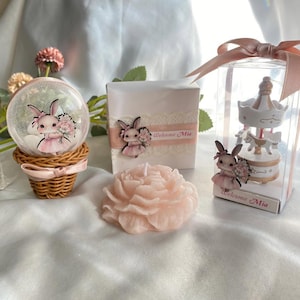 May include: A collection of baby shower favors featuring a snow globe, a gift box, and a carousel figurine. Each item is decorated with a bunny wearing a pink dress and holding flowers. A pink peony-shaped candle is also present. The text "Welcome Mia" is visible.