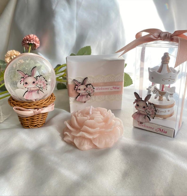 May include: A collection of baby shower favors featuring a snow globe, a gift box, and a carousel figurine. Each item is decorated with a bunny wearing a pink dress and holding flowers. A pink peony-shaped candle is also present. The text "Welcome Mia" is visible.