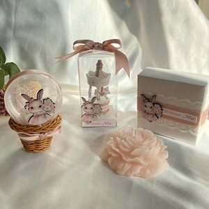 May include: A collection of baby shower favors, including a snow globe, a carousel figurine in a clear box, a gift box, and a peony-shaped candle. Each item features a bunny illustration and the text "Welcome Mia."