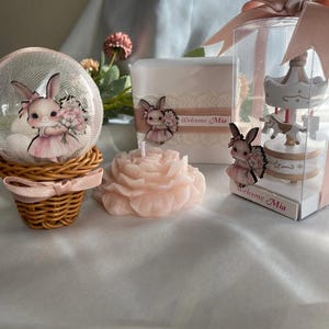 May include: A collection of baby shower decorations featuring a bunny theme. Includes a snow globe, a candle shaped like a rose, and a carousel figurine. The items are in shades of pink and white, with the text "Welcome Mia".