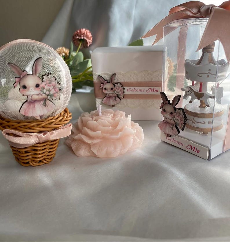 May include: A collection of baby shower decorations featuring a bunny theme. Includes a snow globe, a candle shaped like a rose, and a carousel figurine. The items are in shades of pink and white, with the text "Welcome Mia".