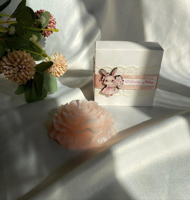 May include: A light pink peony-shaped candle sits next to a white gift box decorated with lace, a pink ribbon, and a bunny figurine. The box has the text "Welcome Mia" on it. Artificial flowers are in the background.