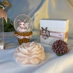 May include: A decorative arrangement featuring a pink peony-shaped candle, a snow globe with a bunny figurine, and a gift box with a bunny and the text "Welcome Mia". The items are in shades of pink and white.