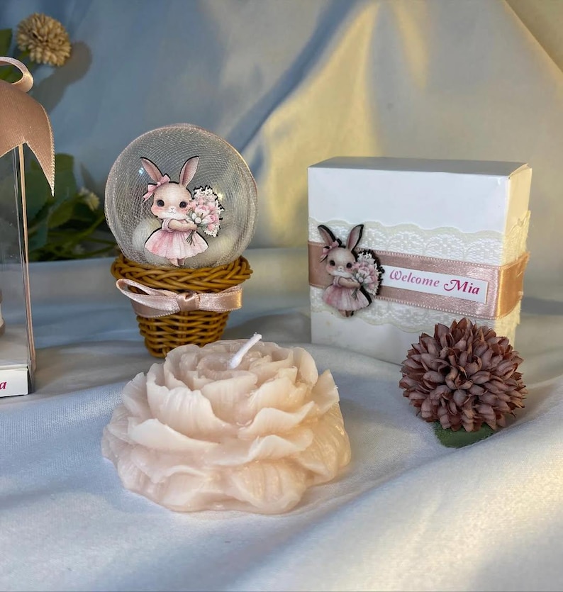 May include: A decorative arrangement featuring a pink peony-shaped candle, a snow globe with a bunny figurine, and a gift box with a bunny and the text "Welcome Mia". The items are in shades of pink and white.