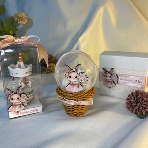 May include: A decorative set featuring a carousel ornament in a clear box, a snow globe with a bunny figure, and a gift box. The bunny figures wear pink dresses and hold bouquets. The text "Welcome Mia" is visible on the box and ornament.