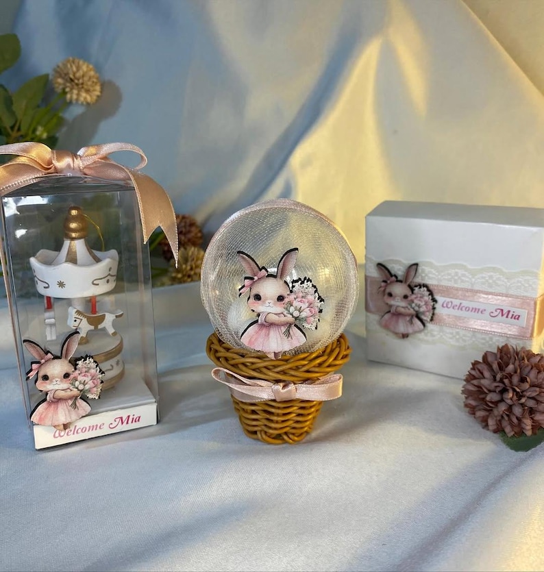 May include: A decorative set featuring a carousel ornament in a clear box, a snow globe with a bunny figure, and a gift box. The bunny figures wear pink dresses and hold bouquets. The text "Welcome Mia" is visible on the box and ornament.