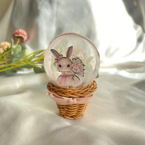 May include: A decorative snow globe featuring a cartoon bunny holding flowers, set inside a clear glass sphere. The globe sits atop a woven basket with a pink ribbon. The bunny wears a pink dress and bow.