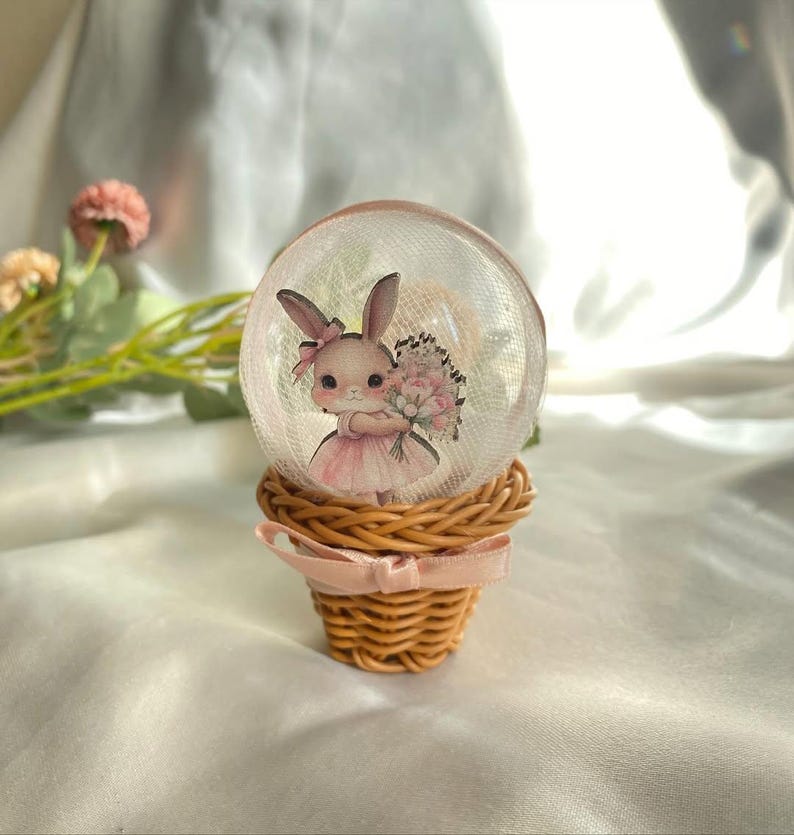 May include: A decorative snow globe featuring a cartoon bunny holding flowers, set inside a clear glass sphere. The globe sits atop a woven basket with a pink ribbon. The bunny wears a pink dress and bow.
