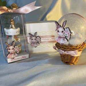 May include: A collection of baby shower favors featuring a carousel, a decorative box, and a snow globe. Each item is adorned with a cartoon bunny holding flowers and the text "Welcome Mia". The favors are decorated with pink ribbons and lace.