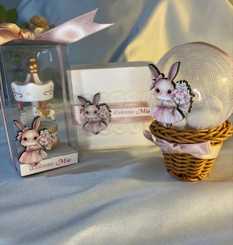 May include: A collection of baby shower favors featuring a carousel, a decorative box, and a snow globe. Each item is adorned with a cartoon bunny holding flowers and the text "Welcome Mia". The favors are decorated with pink ribbons and lace.