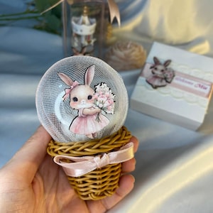 May include: A decorative snow globe featuring a cartoon bunny in a pink dress holding flowers. The globe sits atop a woven basket base tied with a pink ribbon. A small gift box with a bunny image is also visible.