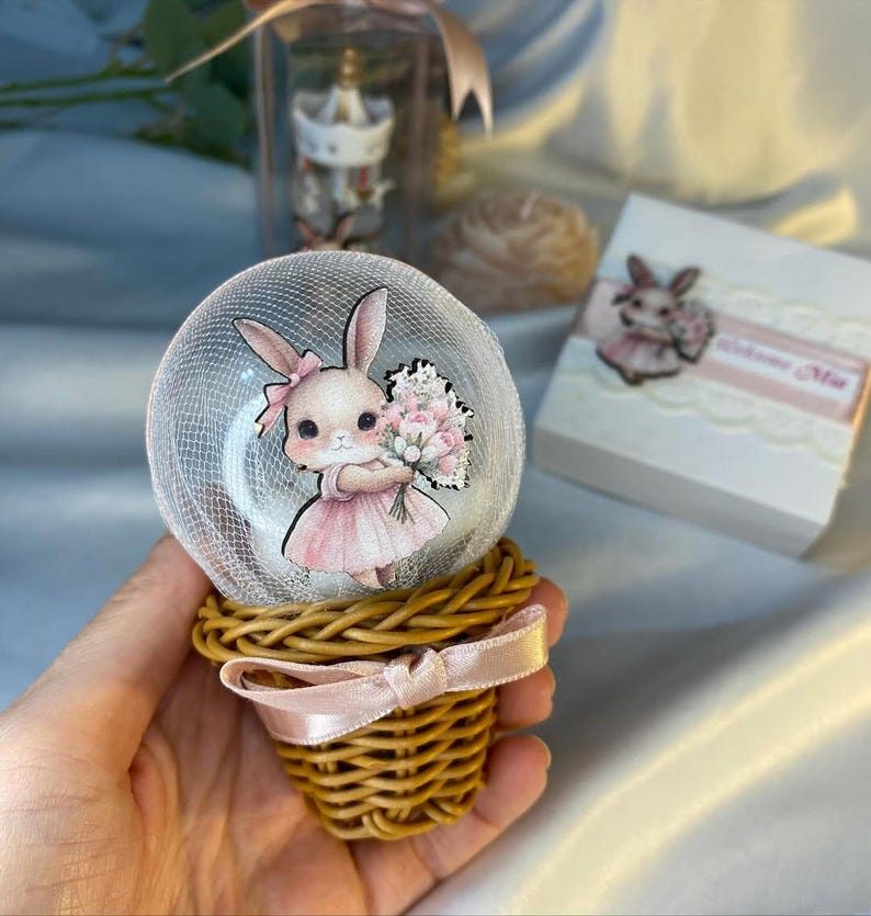May include: A decorative snow globe featuring a cartoon bunny in a pink dress holding flowers. The globe sits atop a woven basket base tied with a pink ribbon. A small gift box with a bunny image is also visible.