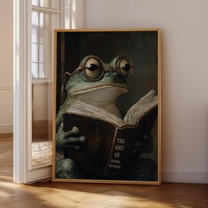 May include: A framed print shows a frog wearing glasses, reading a book titled "The Art of Doing Nothing." The frog is rendered in green and brown tones, holding the book. The artwork is set against a dark background, with a wooden frame.