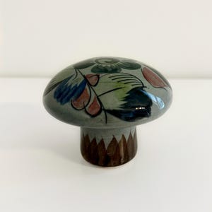 May include: A ceramic mushroom-shaped decorative object with a smooth, glossy finish. The cap is light gray with hand-painted floral motifs in dark green, blue, and reddish-brown. The stem features a brown triangular pattern at the base.