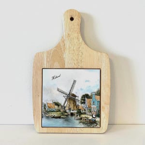 Peut inclure: Planche à découper en bois avec un insert en carrelage rectangulaire représentant un paysage de moulin à vent hollandais. La tuile présente une image peinte d'un moulin à vent, de maisons et d'eau. Le mot "Holland" est écrit sur la tuile. La planche a une poignée avec un trou pour suspendre.