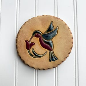 May include: A round, tan-colored ceramic coaster with a hummingbird design. The hummingbird is depicted in shades of burgundy, teal, and green, with a long beak reaching towards a red flower. The coaster has a rough, textured edge.