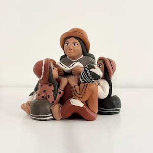May include: A clay figurine set featuring a central figure wearing a brown hat and holding an open book, surrounded by three smaller figures. The figures are painted in earthy tones of brown, black, and white with simple geometric patterns.