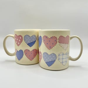 May include: Two cream ceramic mugs featuring heart-shaped patchwork designs in pink, blue, and white. The hearts have various patterns, including polka dots and plaid, stitched onto the mugs.