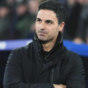 May include: A man with dark hair and a light complexion is wearing a black jacket with a high collar. The jacket has a zipper closure and a quilted lining. The background is blurred, suggesting an outdoor setting.