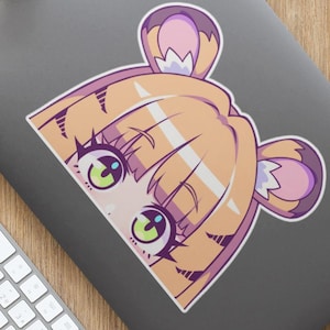 May include: A sticker featuring an anime-style character with light brown hair and purple accents. The character has large, expressive eyes with green irises. The sticker is placed on a gray laptop, surrounded by a keyboard, mouse, headphones, and a coffee mug.