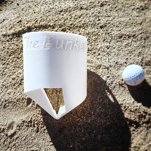 K&ouml;nnte beinhalten: Ein wei&szlig;er Golfball und ein wei&szlig;er Golfballhalter mit dem Text "The Bunker" auf der Seite. Der Halter hat eine dreieckige &Ouml;ffnung an der Unterseite. Die Artikel befinden sich auf einer sandigen Oberfl&auml;che.