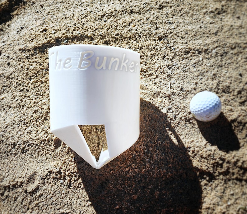 K&ouml;nnte beinhalten: Ein wei&szlig;er Golfball und ein wei&szlig;er Golfballhalter mit dem Text "The Bunker" auf der Seite. Der Halter hat eine dreieckige &Ouml;ffnung an der Unterseite. Die Artikel befinden sich auf einer sandigen Oberfl&auml;che.