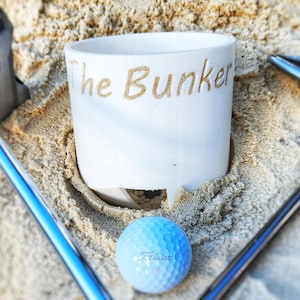 K&ouml;nnte beinhalten: Eine wei&szlig;e Keramiktasse mit dem Schriftzug "The Bunker" in Gold, die in Sand liegt, mit einem blauen Golfball und Golfschl&auml;gern. Der Golfball ist hellblau mit dem Aufdruck "Titleist".