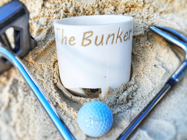 K&ouml;nnte beinhalten: Eine wei&szlig;e Keramiktasse mit dem Schriftzug "The Bunker" in Gold, die in Sand liegt, mit einem blauen Golfball und Golfschl&auml;gern. Der Golfball ist hellblau mit dem Aufdruck "Titleist".