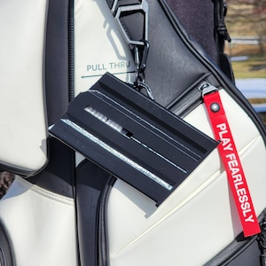 May include: Black golf club accessory with a red tag that says "PLAY FEARLESSLY" attached to a black and white golf bag. The accessory is rectangular with horizontal lines.