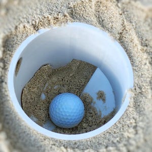 K&ouml;nnte beinhalten: Ein hellblauer Golfball liegt in einem wei&szlig;en Plastikbecher, der mit Sand gef&uuml;llt ist. Der Becher befindet sich in einem sandigen Bereich, wobei der Sand den Becher umgibt. Der Golfball hat eine geriffelte Oberfl&auml;che.