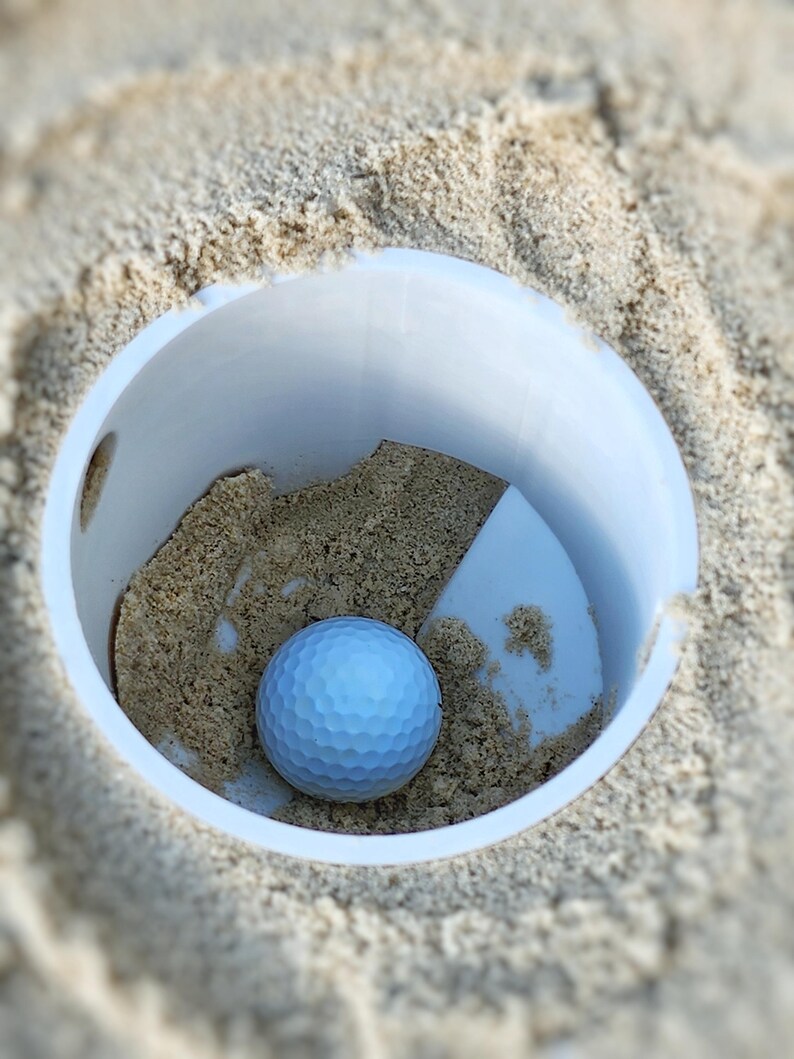 K&ouml;nnte beinhalten: Ein hellblauer Golfball liegt in einem wei&szlig;en Plastikbecher, der mit Sand gef&uuml;llt ist. Der Becher befindet sich in einem sandigen Bereich, wobei der Sand den Becher umgibt. Der Golfball hat eine geriffelte Oberfl&auml;che.