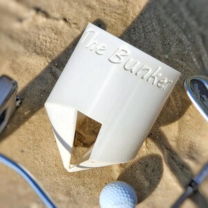 K&ouml;nnte beinhalten: Ein wei&szlig;er Golfballhalter aus Kunststoff mit dem Text "The Bunker" auf der Seite. Der Halter hat eine dreieckige &Ouml;ffnung und ist von Golfschl&auml;gern und einem Golfball auf sandigem Untergrund umgeben.