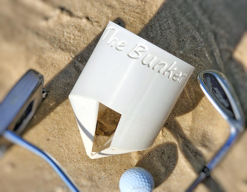K&ouml;nnte beinhalten: Ein wei&szlig;er Golfballhalter aus Kunststoff mit dem Text "The Bunker" auf der Seite. Der Halter hat eine dreieckige &Ouml;ffnung und ist von Golfschl&auml;gern und einem Golfball auf sandigem Untergrund umgeben.
