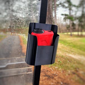 May include: A black golf cart accessory holds a red rangefinder. The holder is mounted on the cart's frame, with a golf course in the background. The rangefinder is a compact, handheld device.
