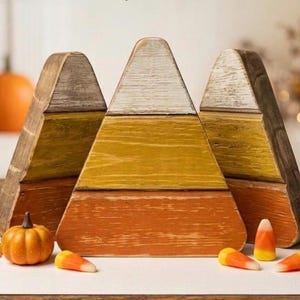 May include: Three triangular wooden decorations shaped like candy corn, featuring horizontal bands of white, yellow, and orange with a distressed finish. The set is arranged with a small artificial pumpkin and several small candy corn pieces.
