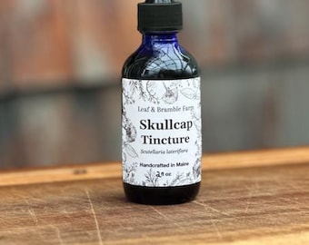 May include: A dark blue glass bottle of Skullcap Tincture with a black dropper. The label features the text "Leaf & Bramble Farm" and "Skullcap Tincture" with botanical illustrations. The bottle contains 2 fl oz of liquid.