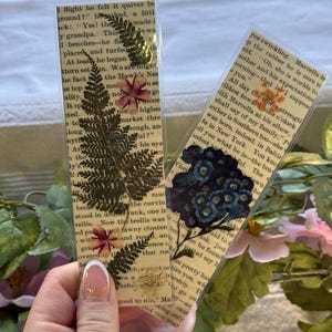 May include: Two handmade bookmarks with pressed flowers and greenery. One bookmark features a fern and small pink flowers, the other has blue flowers. Both are set against a backdrop of vintage book pages.