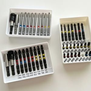 Könnte beinhalten: Drei weiße Kunststoffschalen mit verschiedenen Metall-Schraubendreher-Bits. Die Bits verfügen über farbcodierte Ringe und aufgedruckte Beschriftungen wie PH1, PH2, PZ1, PZ2, T15, T20, T25, T30 und T40, die verschiedene Größen und Antriebstypen angeben.