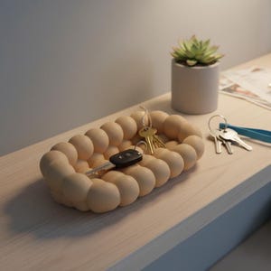 May include: A beige, bubble-shaped key holder containing a car key and other keys. A small succulent in a concrete pot and a set of keys with a blue tag are also on a light wood surface.