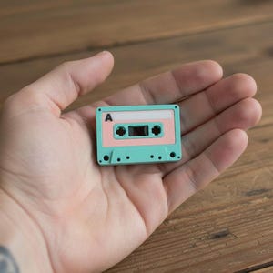 Può includere: Una piccola cassetta audio turchese e rosa con la lettera "A" stampata sopra. La cassetta è tenuta in una mano. Lo sfondo è una superficie di legno.