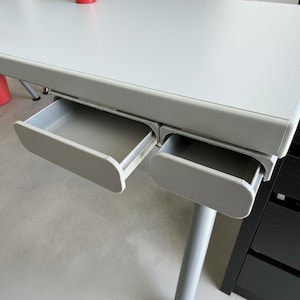 Può includere: Una scrivania grigio chiaro con due cassetti aperti. I cassetti sono rettangolari con bordi arrotondati e sono incassati nel telaio della scrivania. La scrivania ha un design minimalista ed è sostenuta da un'unica gamba cilindrica.