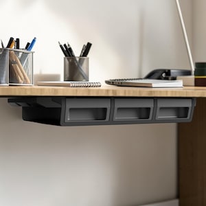 Puede incluir: Un escritorio de madera con una unidad de cajones negra debajo del escritorio. La unidad tiene tres cajones. Varios útiles de oficina, incluidos bolígrafos, lápices y cuadernos, están sobre el escritorio. El escritorio está contra una pared blanca.